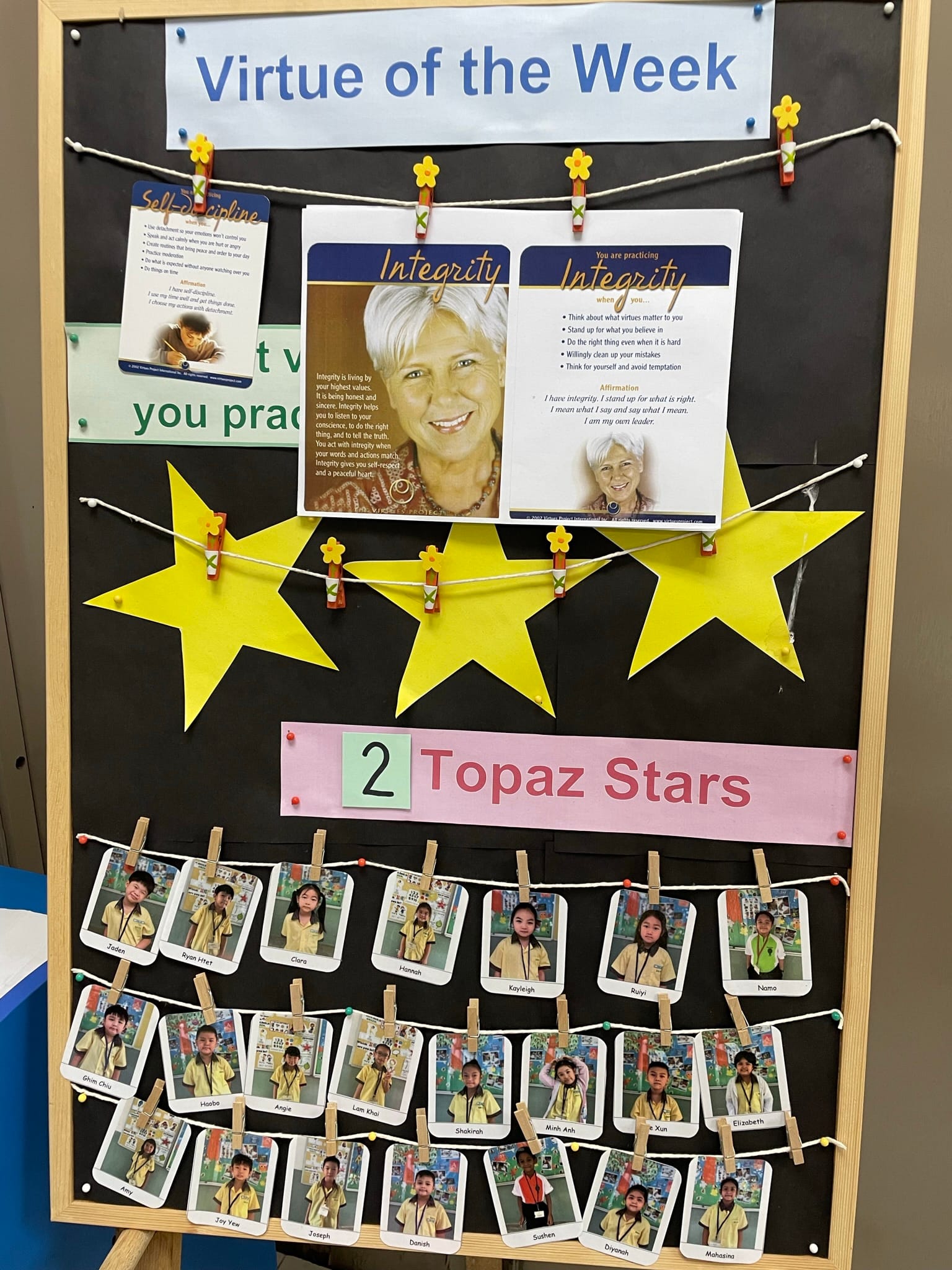 A classroom bulletin board titled "Virtue of the Week" featuring Integrity cards, yellow stars, and student photos labeled "2 Topaz Stars."